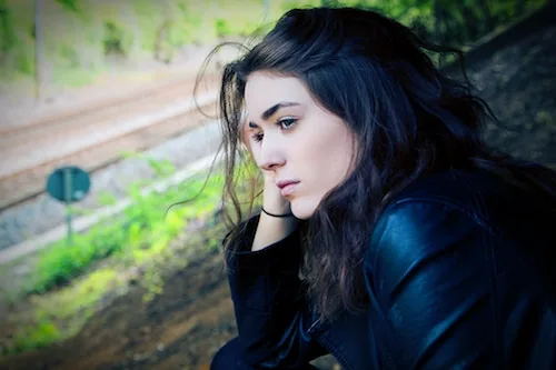 Woman with dark hair in profile, wearing dark jacket, resting head on hand with contemplative expression outdoors